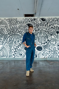 Matteo Miles, mural artist, standing on concrete floor in coveralls before one of his murals in black and white, depicting abstracted images of fingers, eyes and hands.