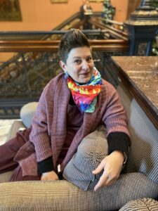 A white woman with short spikey brown hair wears a rainbow scarf while sitting on a couch in a retro lobby.
