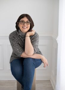 A white woman with chin-length dark brown hair and glasses sits cross-legged in a grey turtleneck against a white wall.